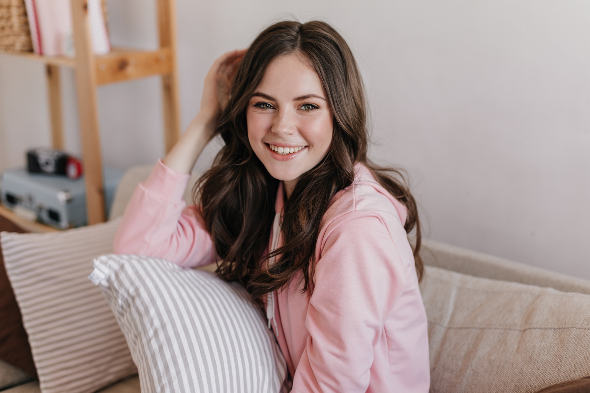 Portrait photo at home. Beautiful brunette girl dressed in pink sweatshirt brightly smiling and pos